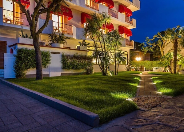L’extérieur d’un hôtel, bien éclairé la nuit, avec un trottoir dallé, une pelouse soigneusement entretenue et des palmiers.