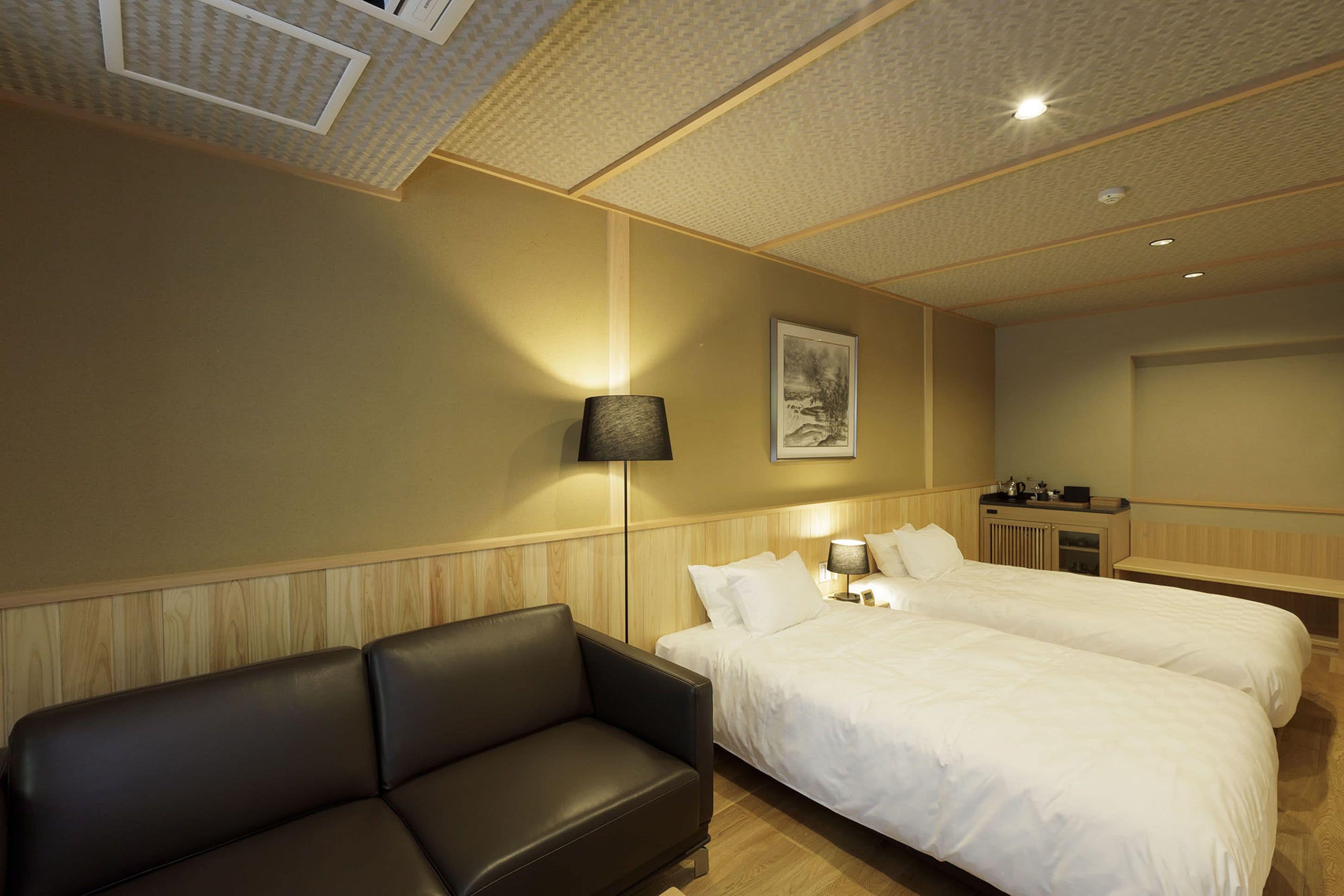 Een hotelkamer met een houten hoofdbord, twee bedden met witte lakens, een zwarte leren bank, een nachtkastje met een lamp en een ingelijst schilderij aan de muur.