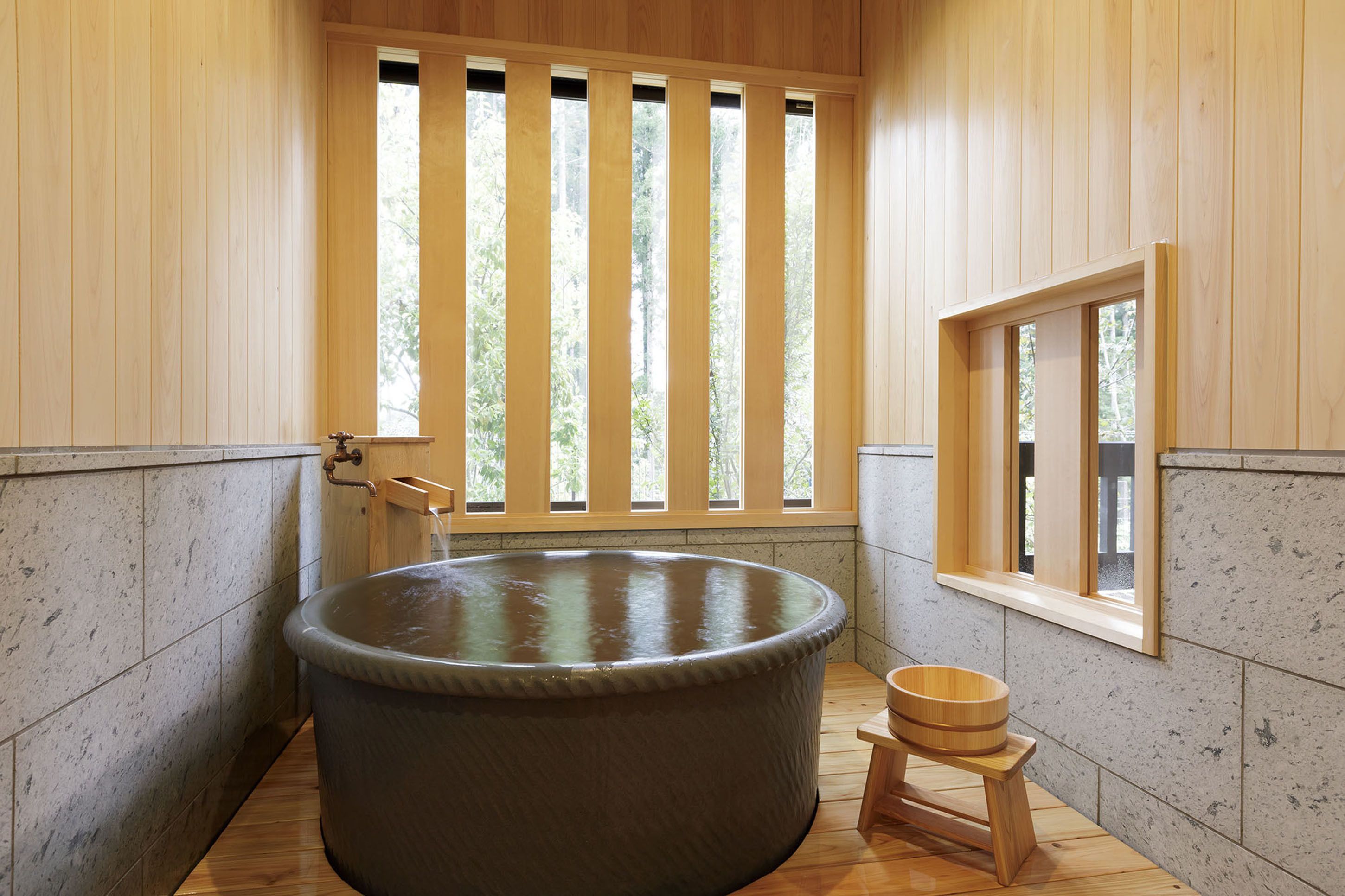 A wooden spa room with a large hot tub, a wooden stool, and a window with vertical blinds.