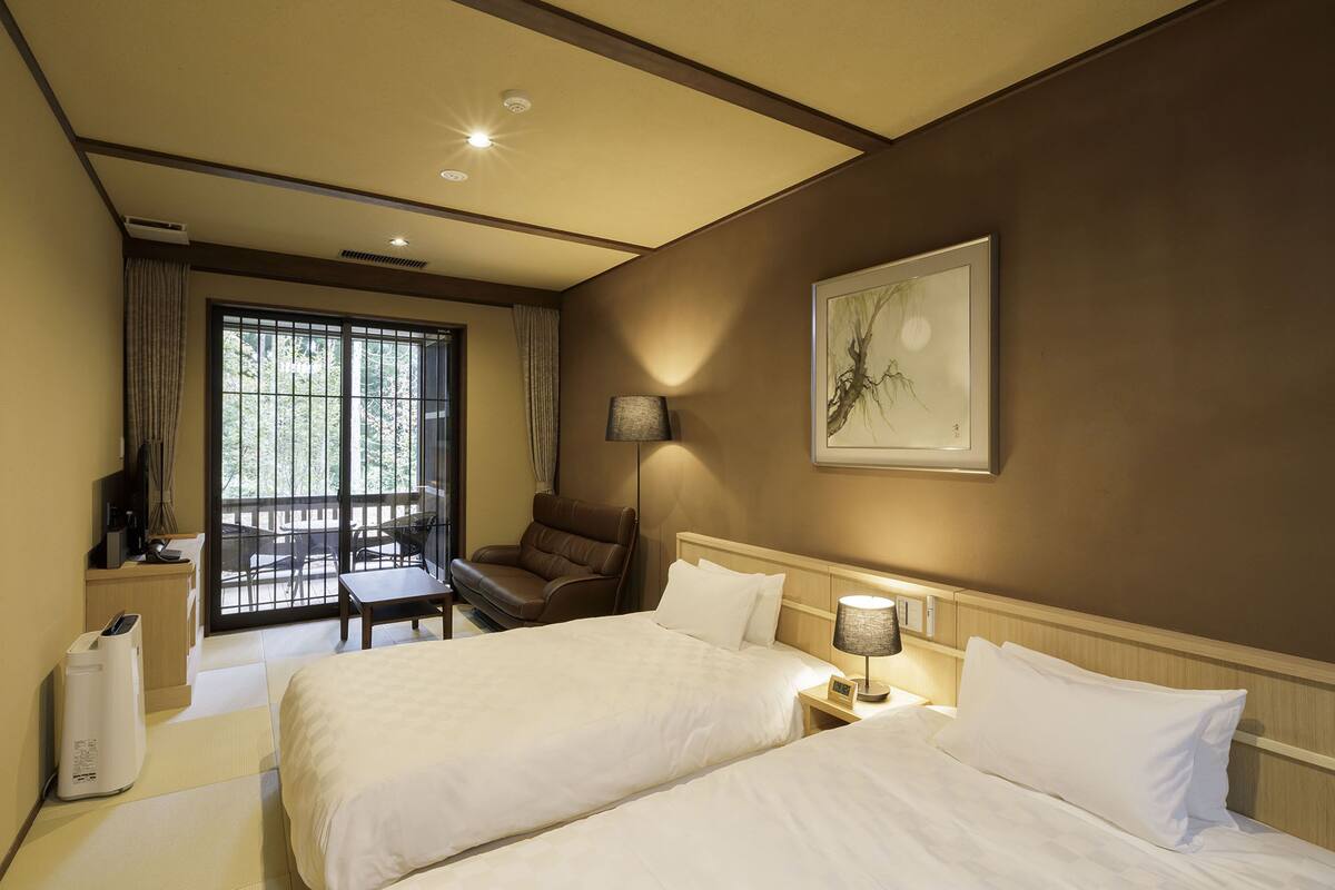 Une chambre d’hôtel avec deux lits, un bureau avec une chaise, un canapé, une grande fenêtre et un tableau au mur.