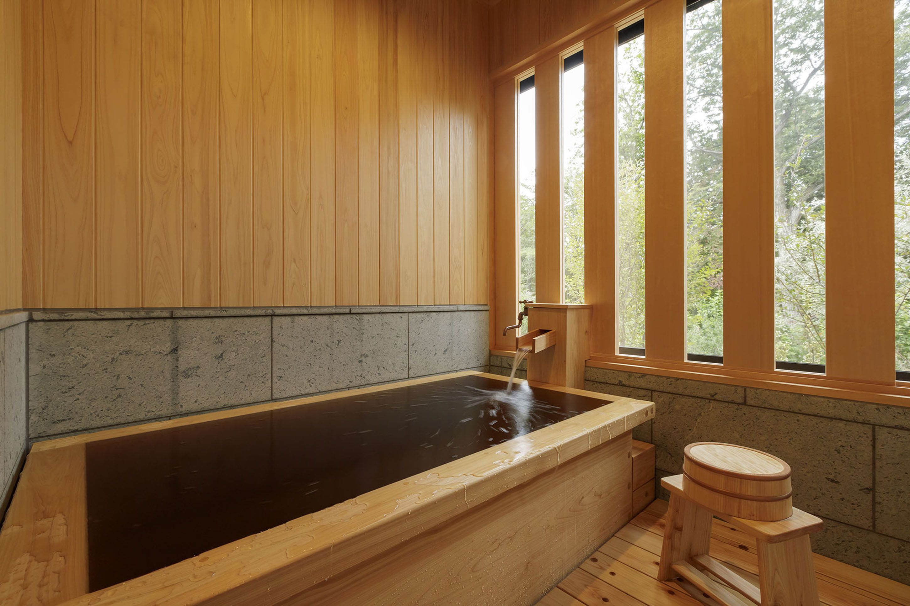 A wooden spa bath with a stone wall, a wooden bench, and a window with a view of trees.