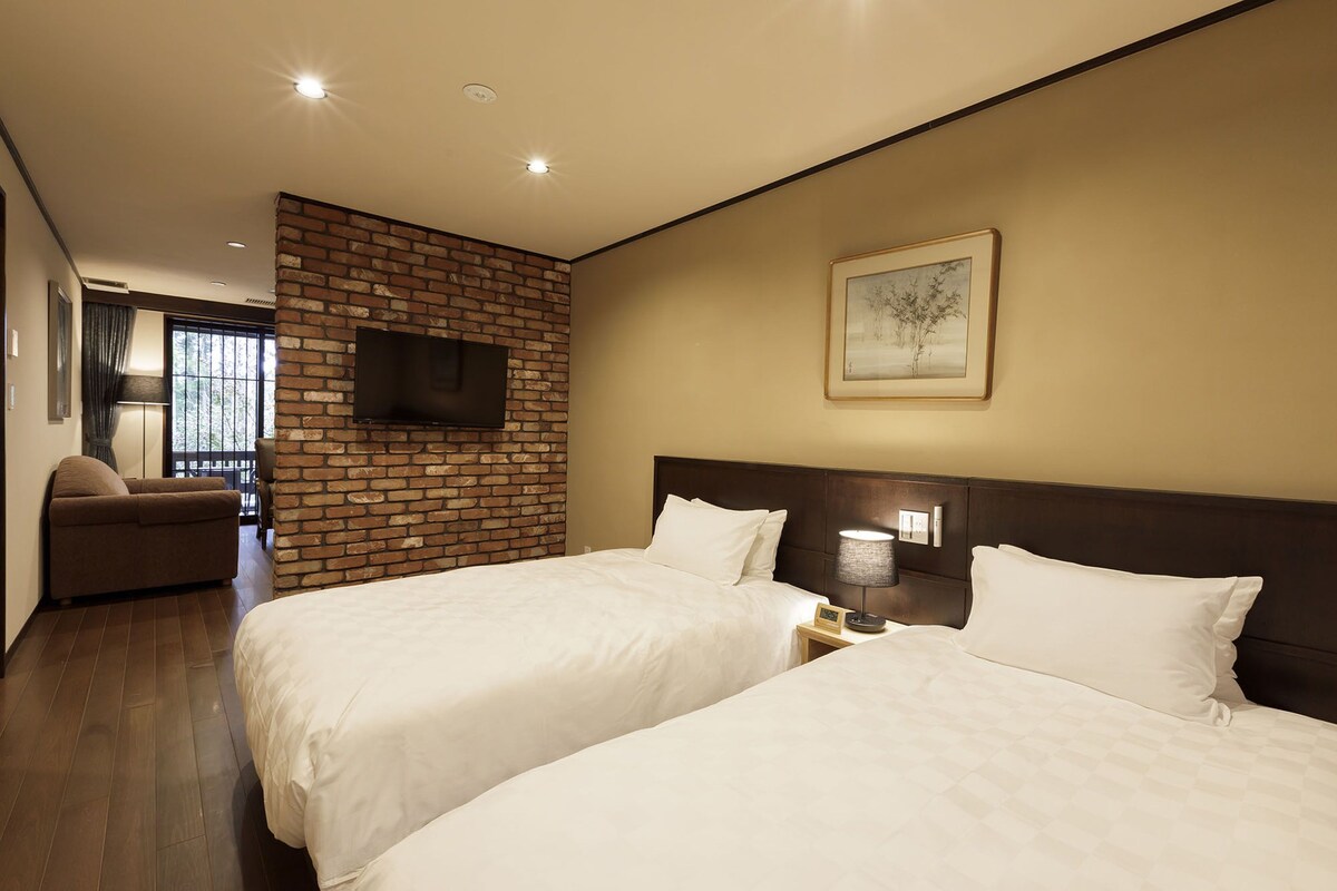 A hotel room with two beds, a brick accent wall, a flat-screen TV mounted above the bed, a framed artwork on the wall, and a window with curtains.