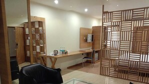 Habitación de hotel moderna con una mesa de centro de vidrio, sofá de cuero negro, separador de madera y un escritorio con computadora y botellas de agua.