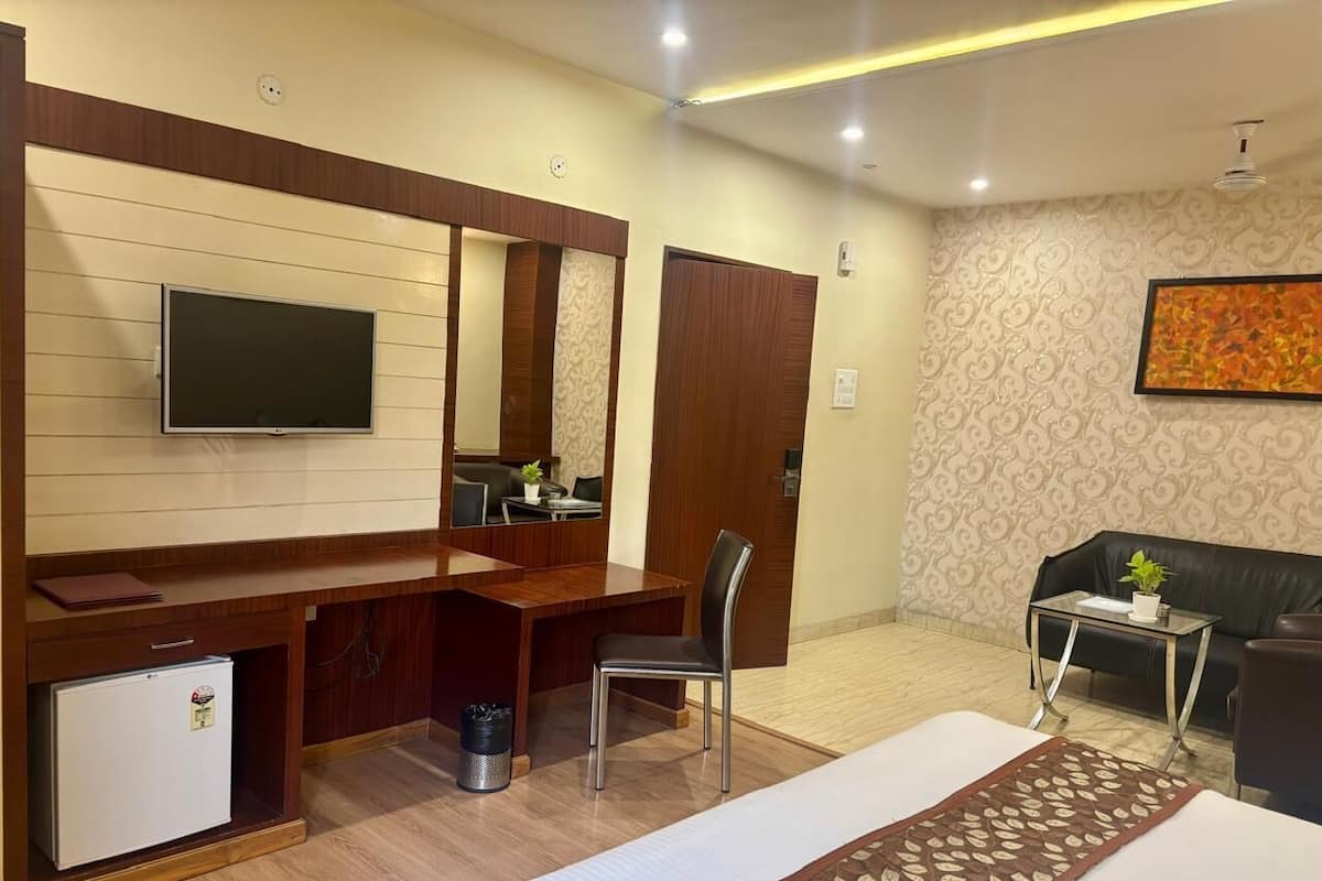 A hotel room with a bed, a desk, a chair, a small refrigerator, a trash bin, a sofa, a coffee table, and a wall-mounted TV.