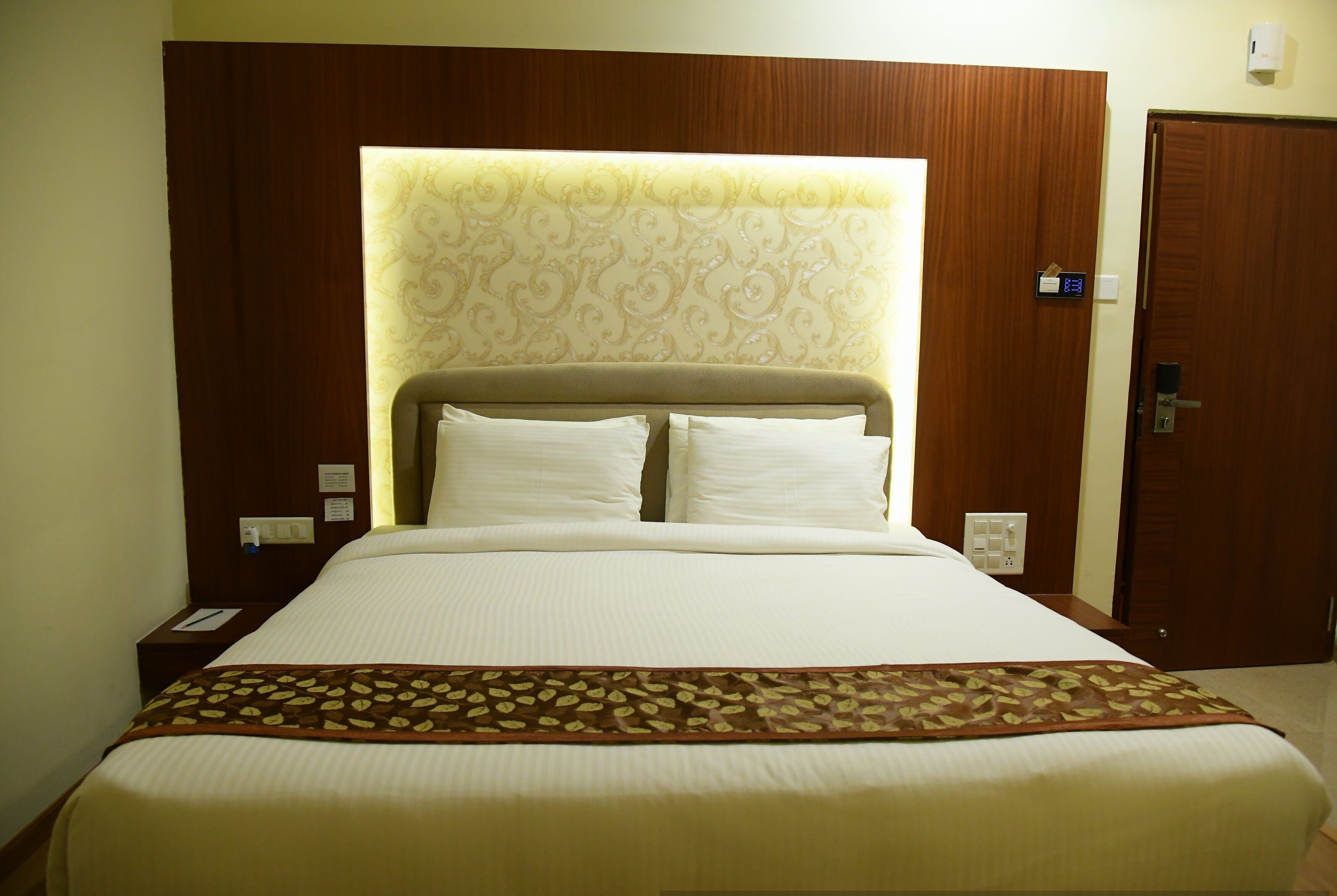 Ein Hotelzimmer mit einem großen Bett, einem Kopfteil mit Muster, einem Nachttisch mit Lampe und einer Tür mit Schloss.