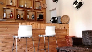 A bar area with wooden shelves displaying bottles and glasses, a coffee machine, and two white bar stools. - Hotel Riva (Rimini)