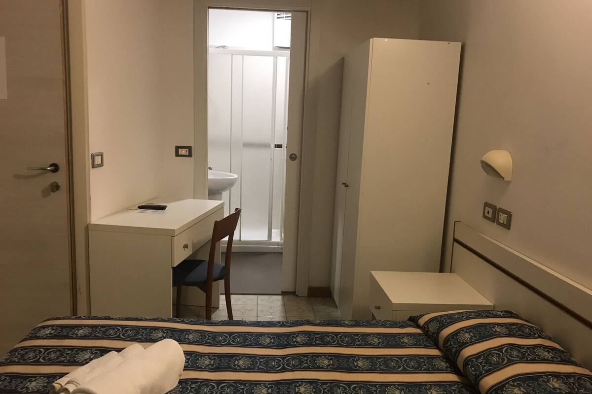 A compact hotel room with a bed, a desk, a chair, a wardrobe, and a bathroom visible through an open door.