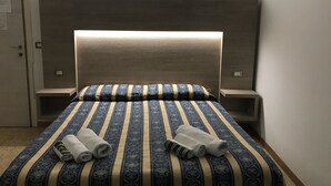 A neatly made bed with striped bedding and two rolled towels, a bedside table, and a wall-mounted light. - Hotel Riva (Rimini)