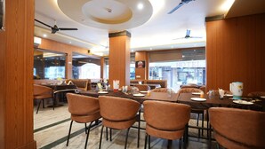 A restaurant with a long communal table, brown upholstered chairs, ceiling fans, and wooden paneling. - HOTEL MADURAI (Udaipur)