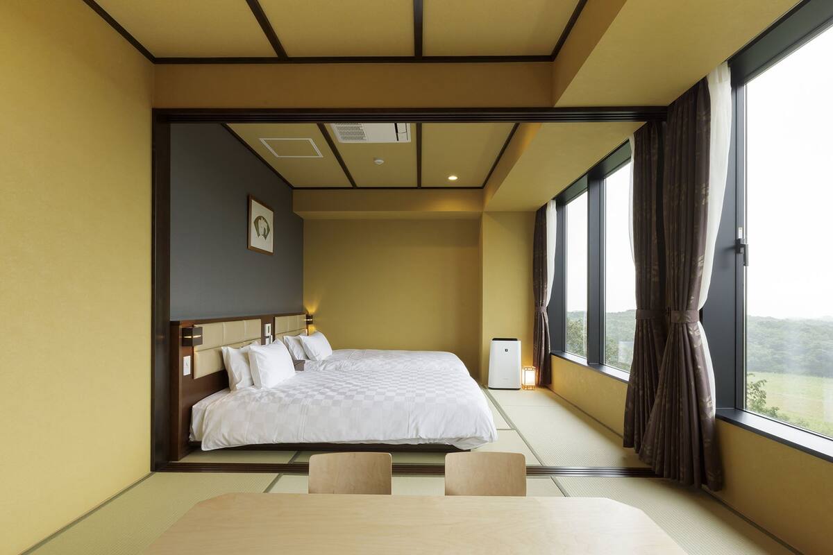 Habitación de hotel con una cama grande, una mesita de noche y vistas al exterior a través de amplias ventanas.