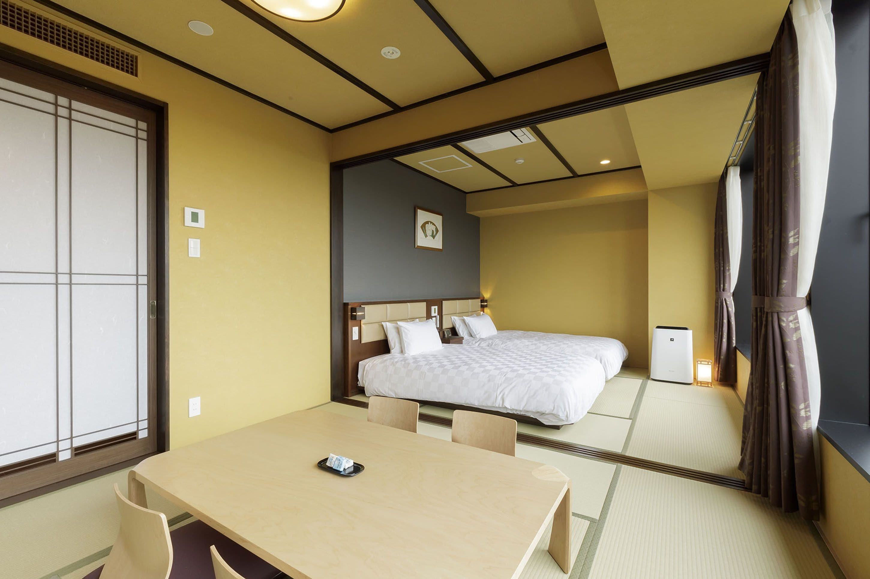 A hotel room with a bed, a bedside table, a chair, a window with a view, and a mirror reflecting the room.