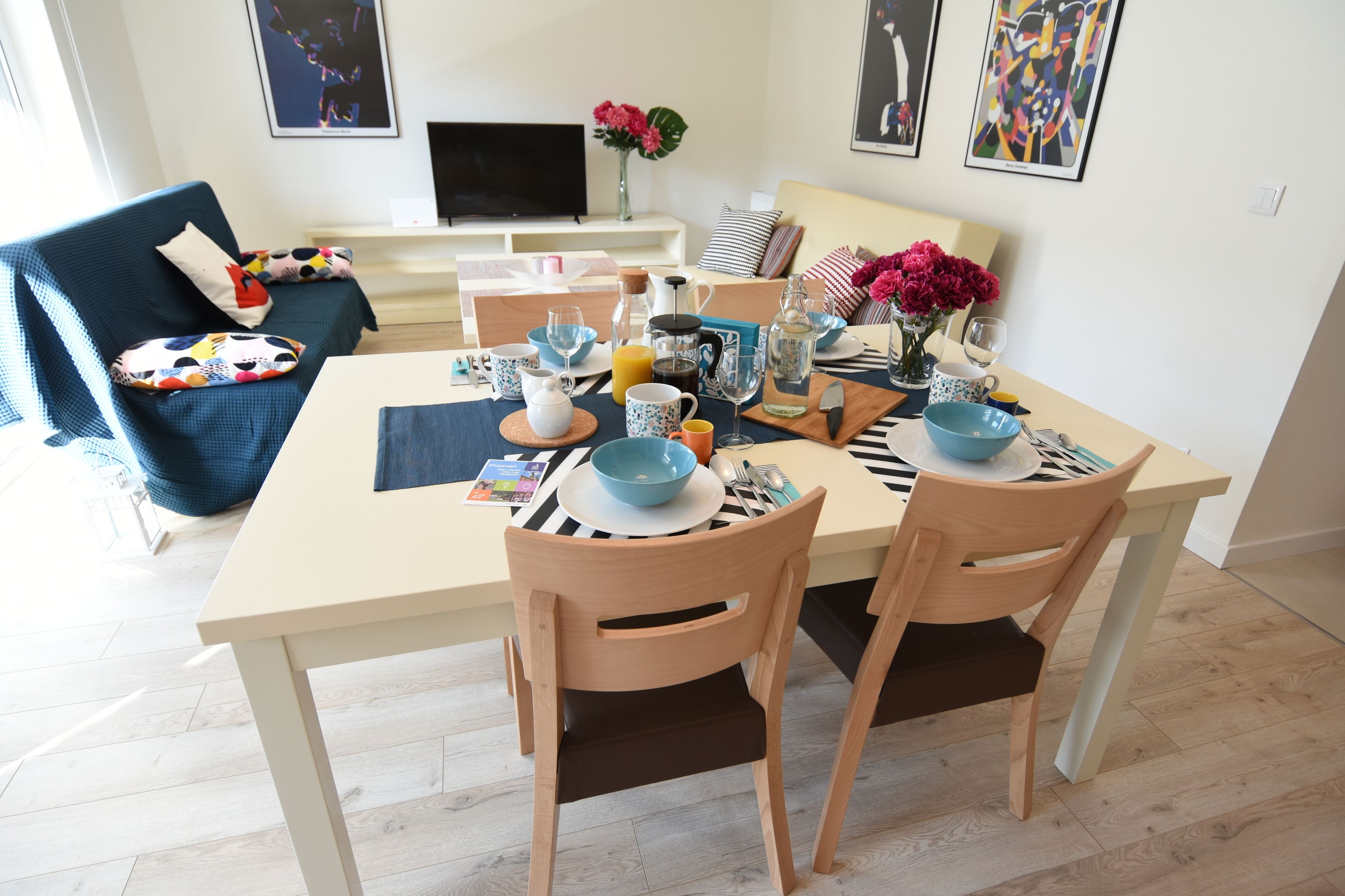 A dining area with a table set for two, featuring a bowl, teapot, and various cups. A sofa and a TV are visible in the background.