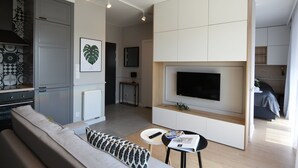 Ein modernes Wohnzimmer mit einer Couch, einem Couchtisch, einem an der Wand befestigten Fernseher und einer Küchenzeile mit Schränken und einer Arbeitsplatte.