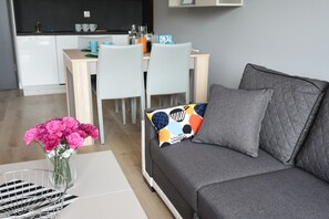 A modern living room with a grey sofa, a coffee table with a vase of pink flowers, and a dining area in the background.