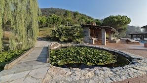 Un sendero de piedra que lleva a un estanque con lirios, rodeado de vegetación y un edificio de piedra con un patio cubierto.