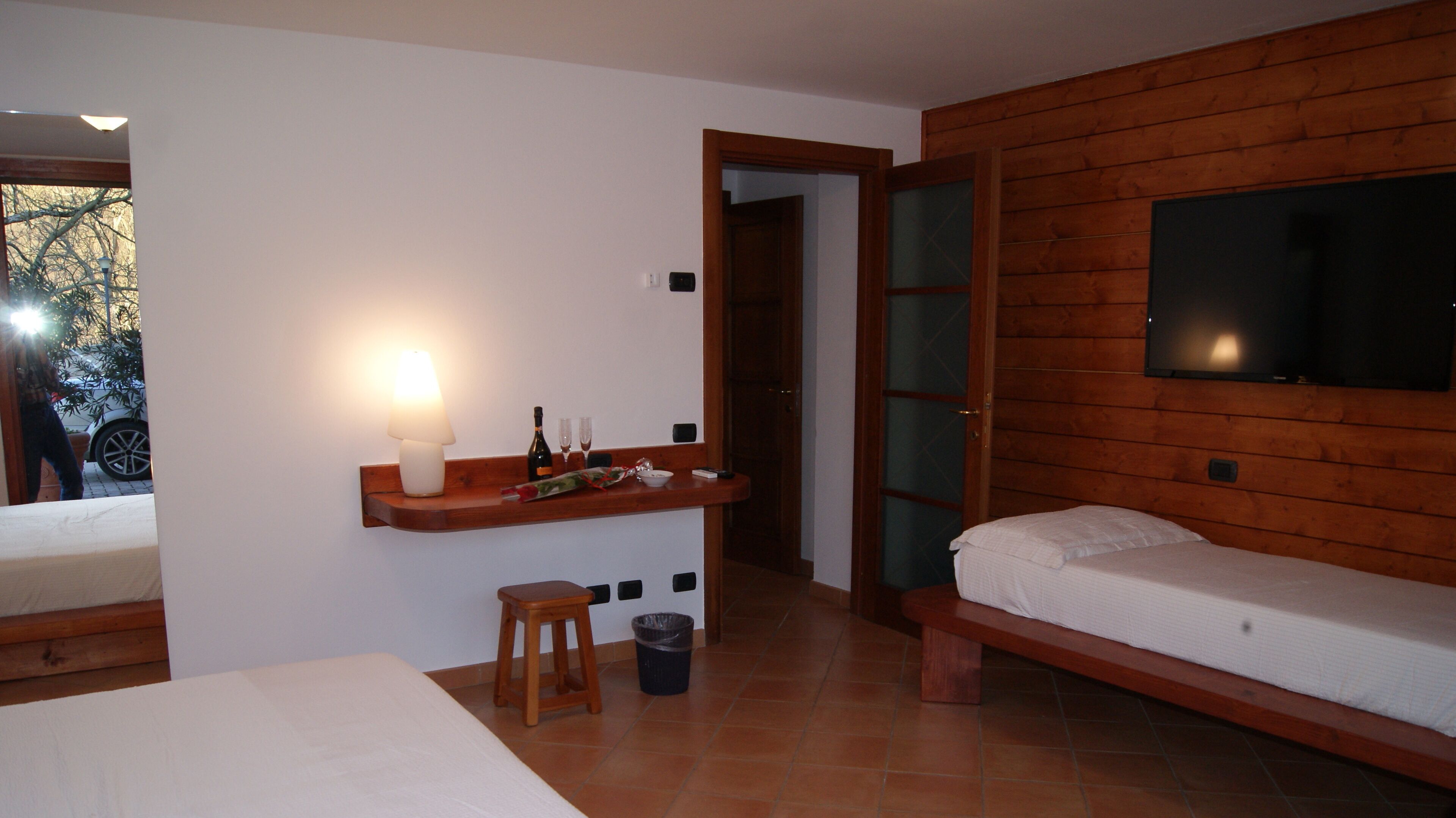A bedroom with a bed, a small stool, a nightstand with a lamp, a TV mounted on the wall, and a wooden door.