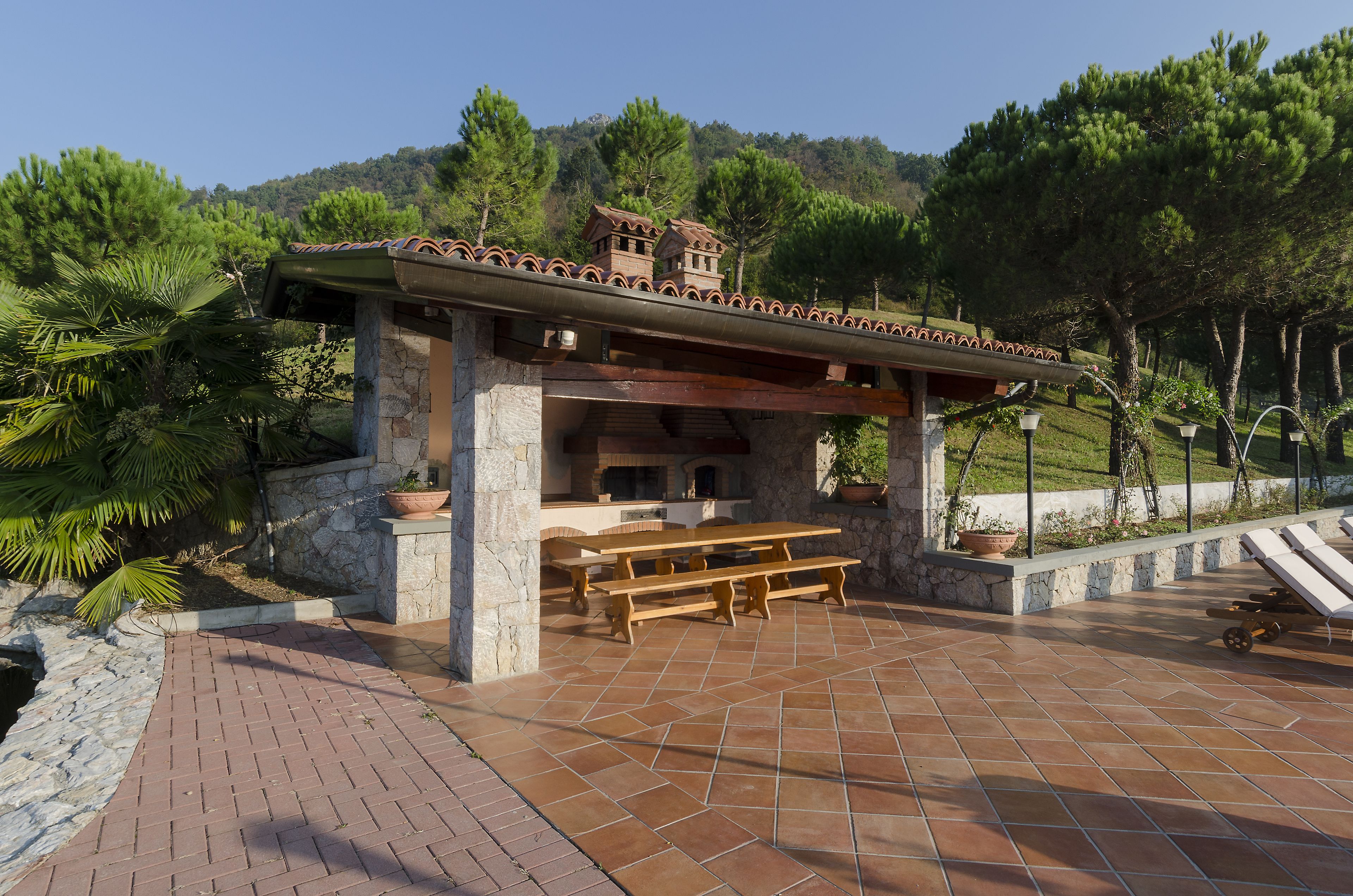 A stone patio with a wooden table and benches, a small brick structure with a tiled roof, and a view of a hillside with trees.