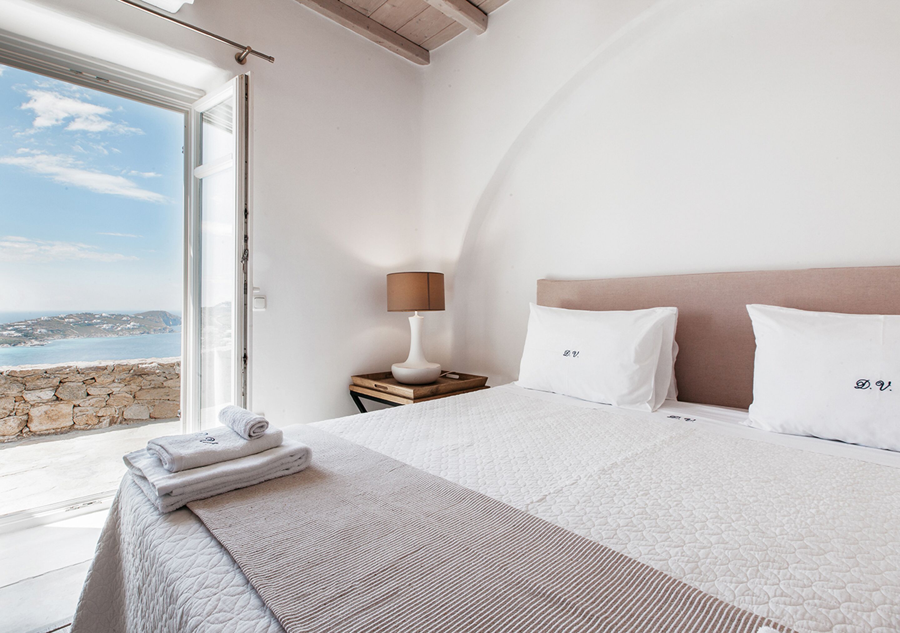 Une chambre à coucher avec un grand lit, une table de chevet avec une lampe, et une vue sur la mer par la fenêtre.