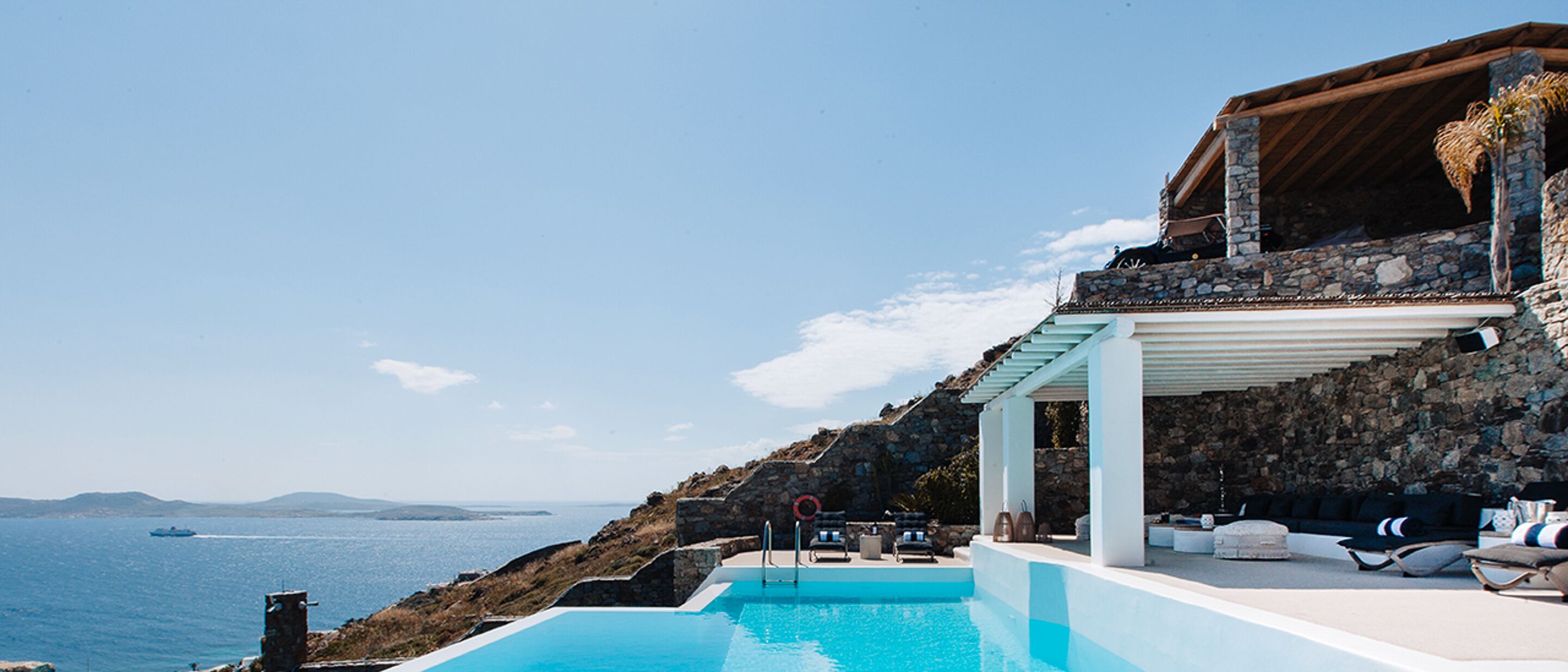 Un espace aménagé au bord de la piscine, avec vue sur la mer, des murs en pierre et des sièges d’extérieur.
