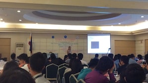 Une salle de conférence où les participants sont assis à des tables, avec un écran de présentation et un drapeau en arrière-plan.