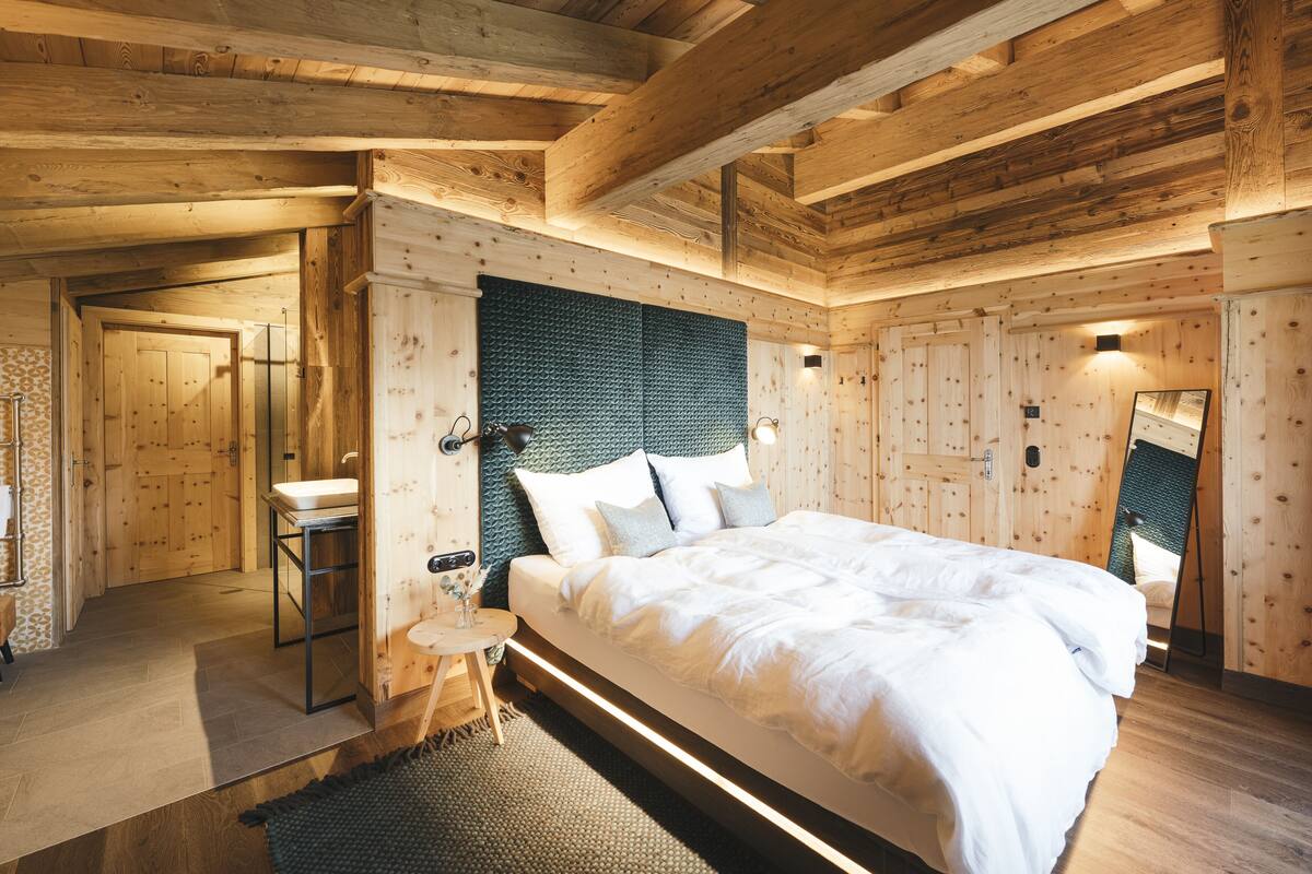A wooden bedroom with a large bed, a small bedside table, a mirror, and a door leading to a bathroom.