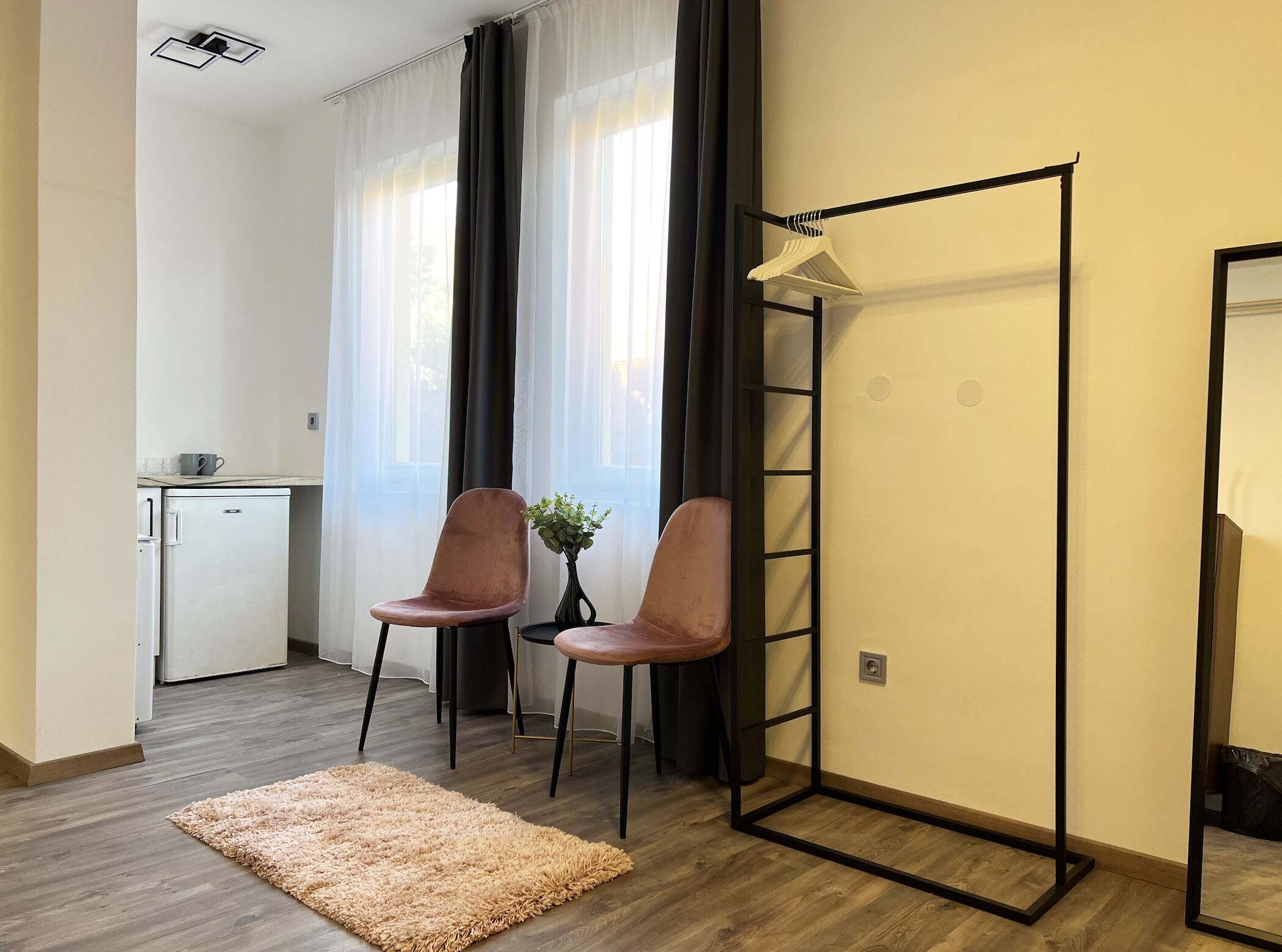 A compact room with a kitchenette, two chairs, a small table, a mirror, and a large window with sheer curtains.