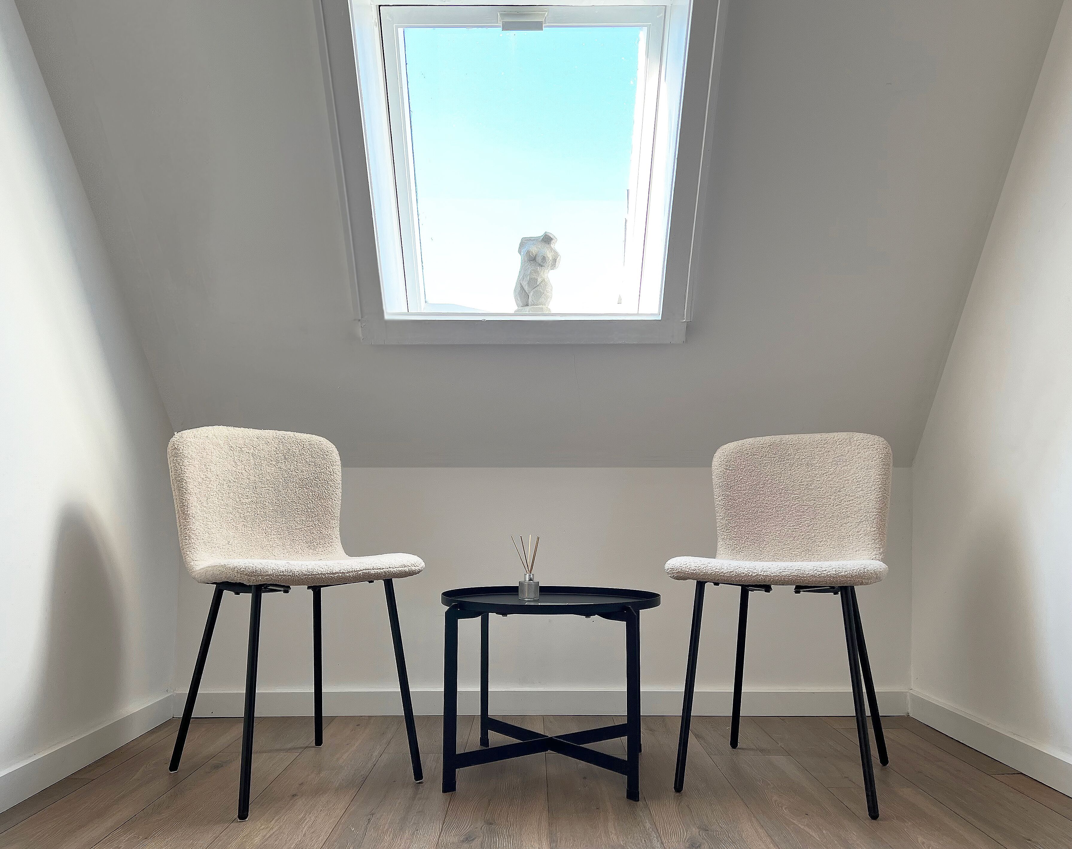 Una habitación pequeña con dos sillas y una mesa, una ventana con vistas, y suelo de madera.