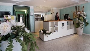 A reception desk with a receptionist, a large potted plant, and a mirror reflecting the interior. - Si Rimini Hotel (Rimini)