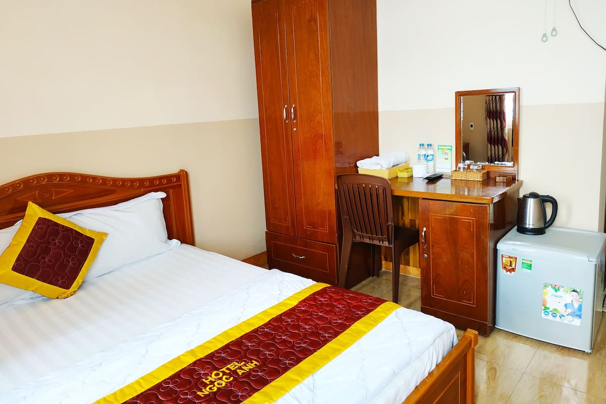 A hotel room with a bed, a wooden wardrobe, a desk with a chair, a small refrigerator, and a mirror.