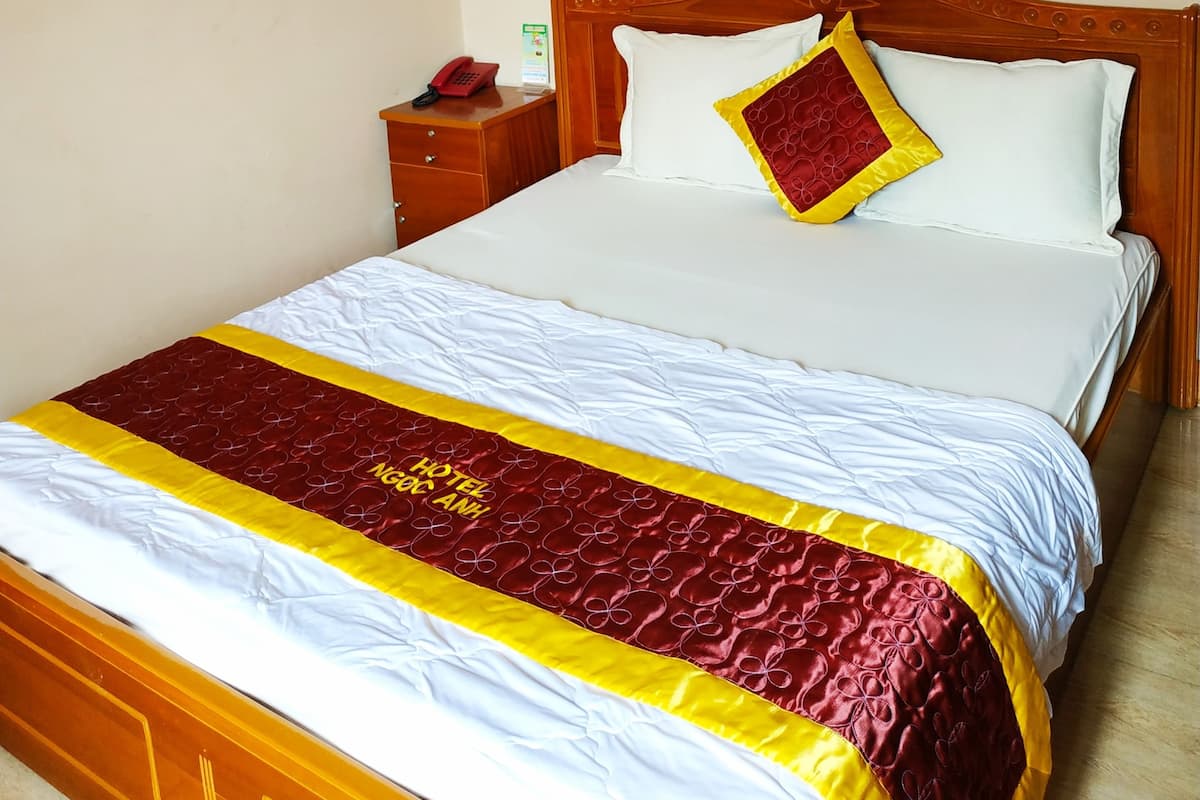 A hotel room with a wooden bed, white bedding, a red and yellow bedspread, a bedside table with a telephone, and a wall socket.