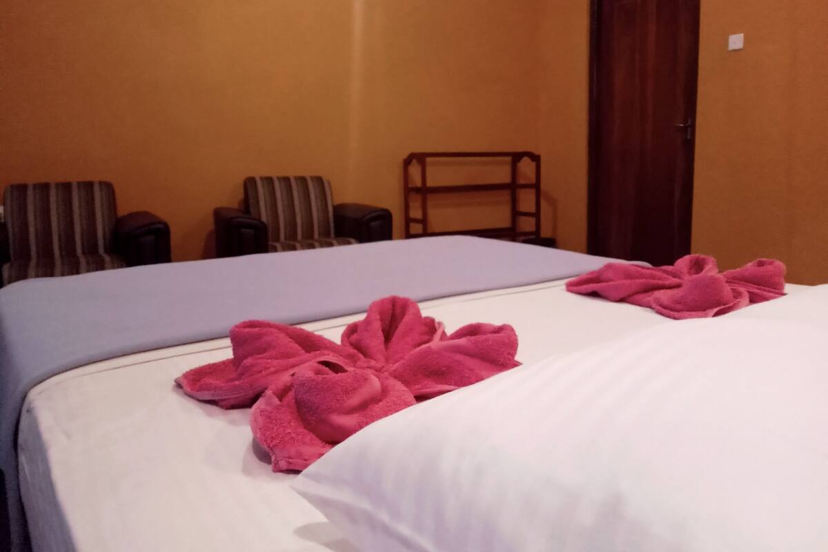 Two beds with white and blue bedding, pink towels, and a wooden chair in a room with orange walls.