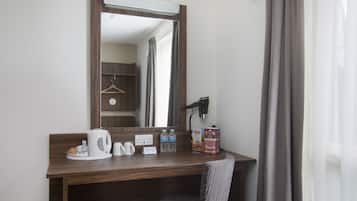 Habitación de hotel con un escritorio de madera, un espejo, una silla, un hervidor, una cafetera, un secador de pelo y un armario con perchas.