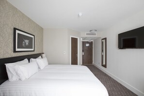 A hotel room with a large bed, a mounted TV, a framed picture on the wall, and a door leading to another room.