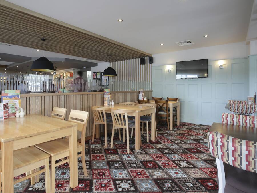 Een restaurant met houten tafels en stoelen, een vloerkleed met patroon en een aan de muur bevestigde televisie.