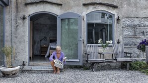 A woman reading a newspaper outside a building with a stone facade, a wooden bench, and a potted plant. - Fårösunds Fästning (Fårösund)