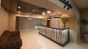 Un área de recepción con diseño moderno, que presenta un mostrador con encimera de vidrio y una planta decorativa. A la izquierda, un sofá marrón y, a la derecha, una planta grande en maceta.