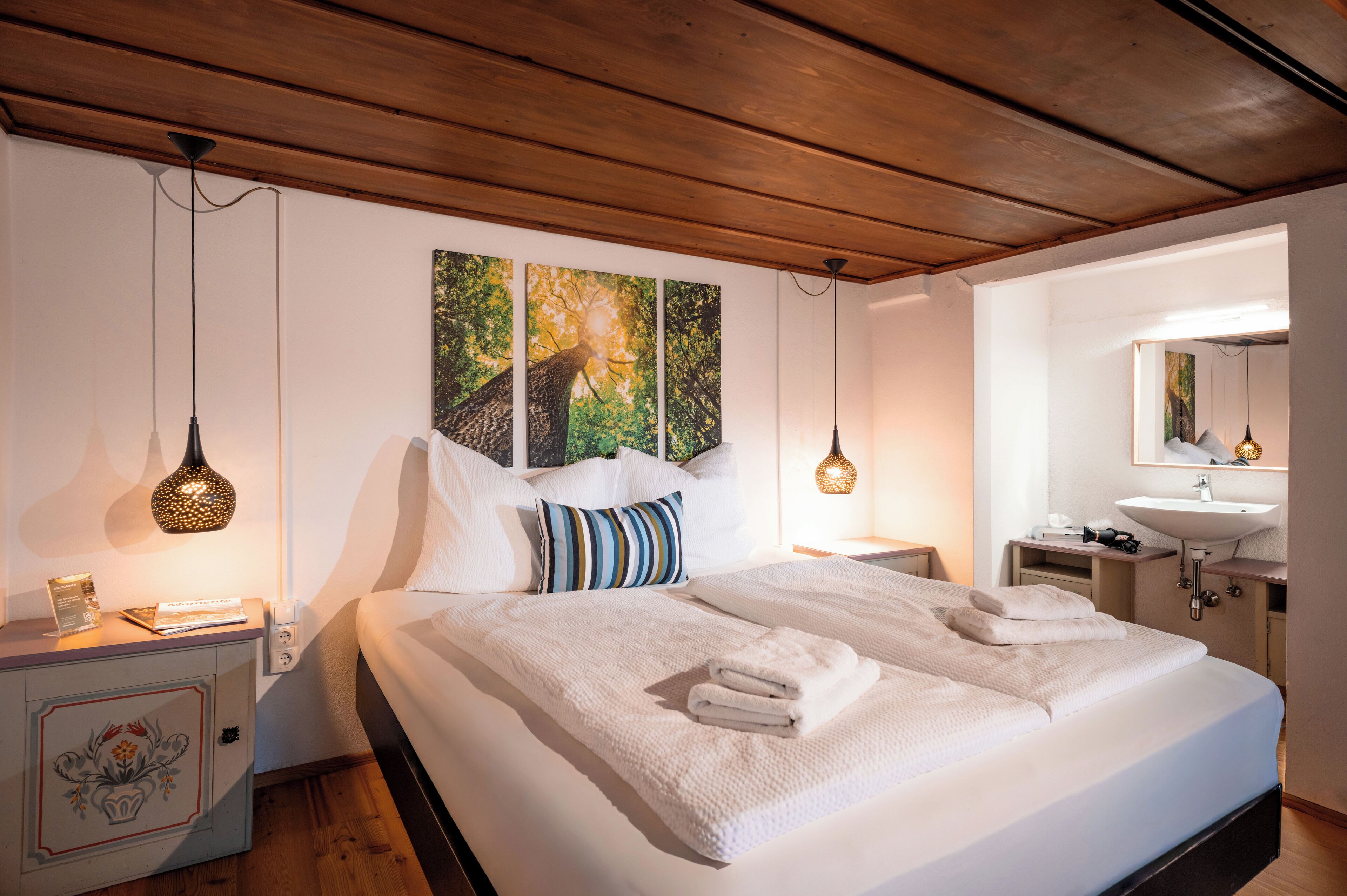 Une chambre d’hôtel avec un grand lit, une table de chevet avec une lampe, une commode, un plafond en bois et un miroir fixé au mur.