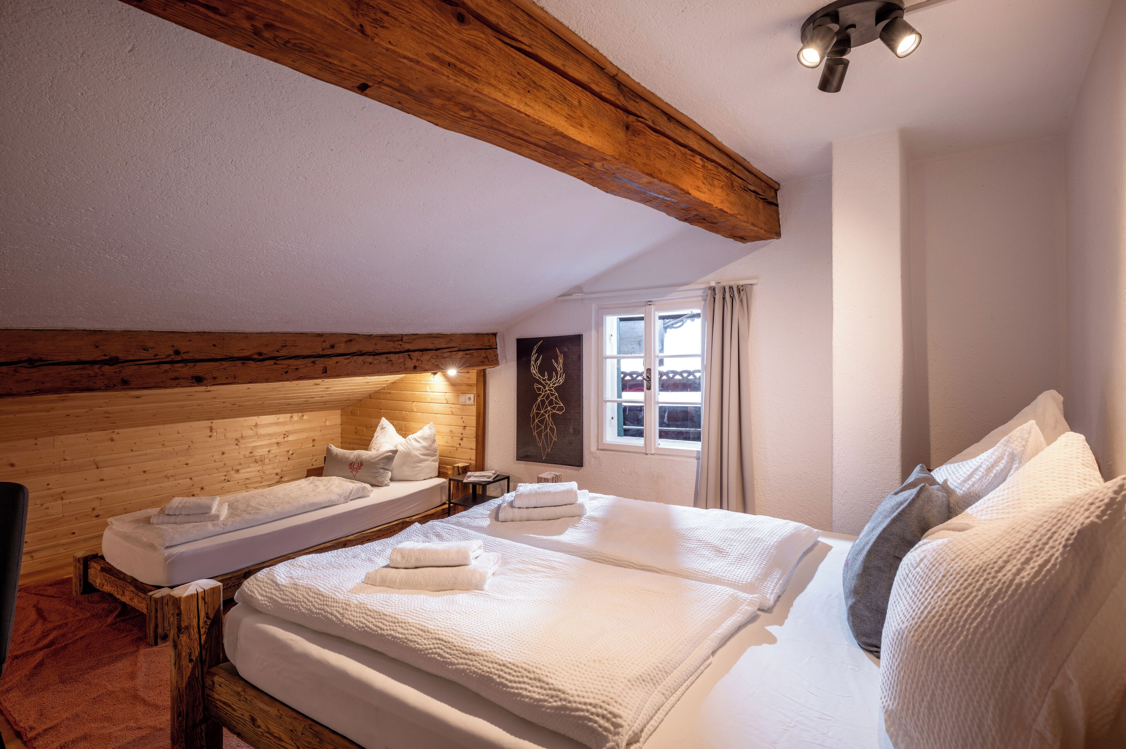 A cozy room with two beds, wooden beams, a window with curtains, and a ceiling light fixture.