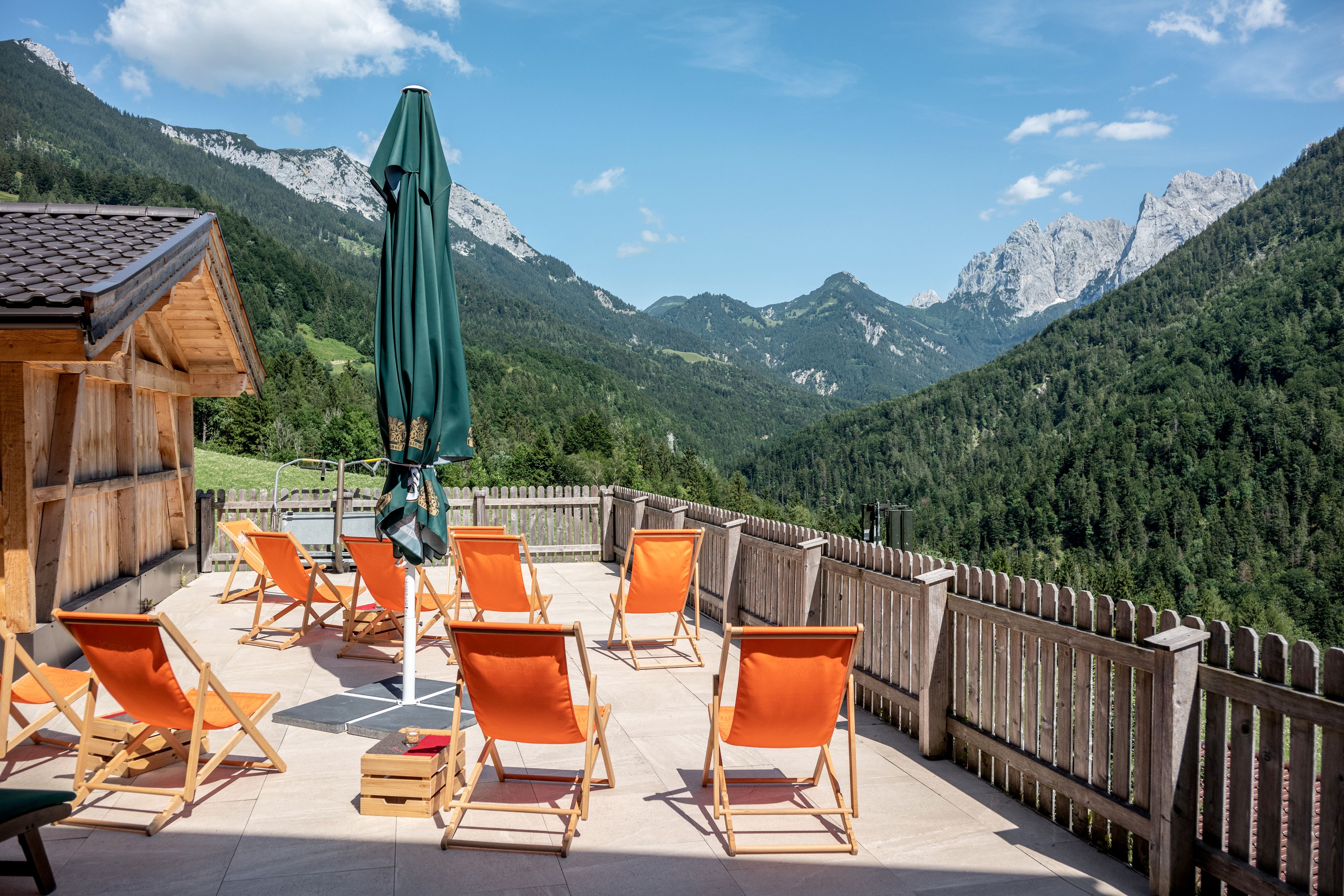 Een houten terras met oranje stoelen en een groene parasol, met uitzicht op een bergachtig landschap.