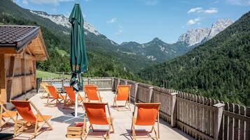 Een houten terras met oranje stoelen en een groene parasol, met uitzicht op een bergachtig landschap.