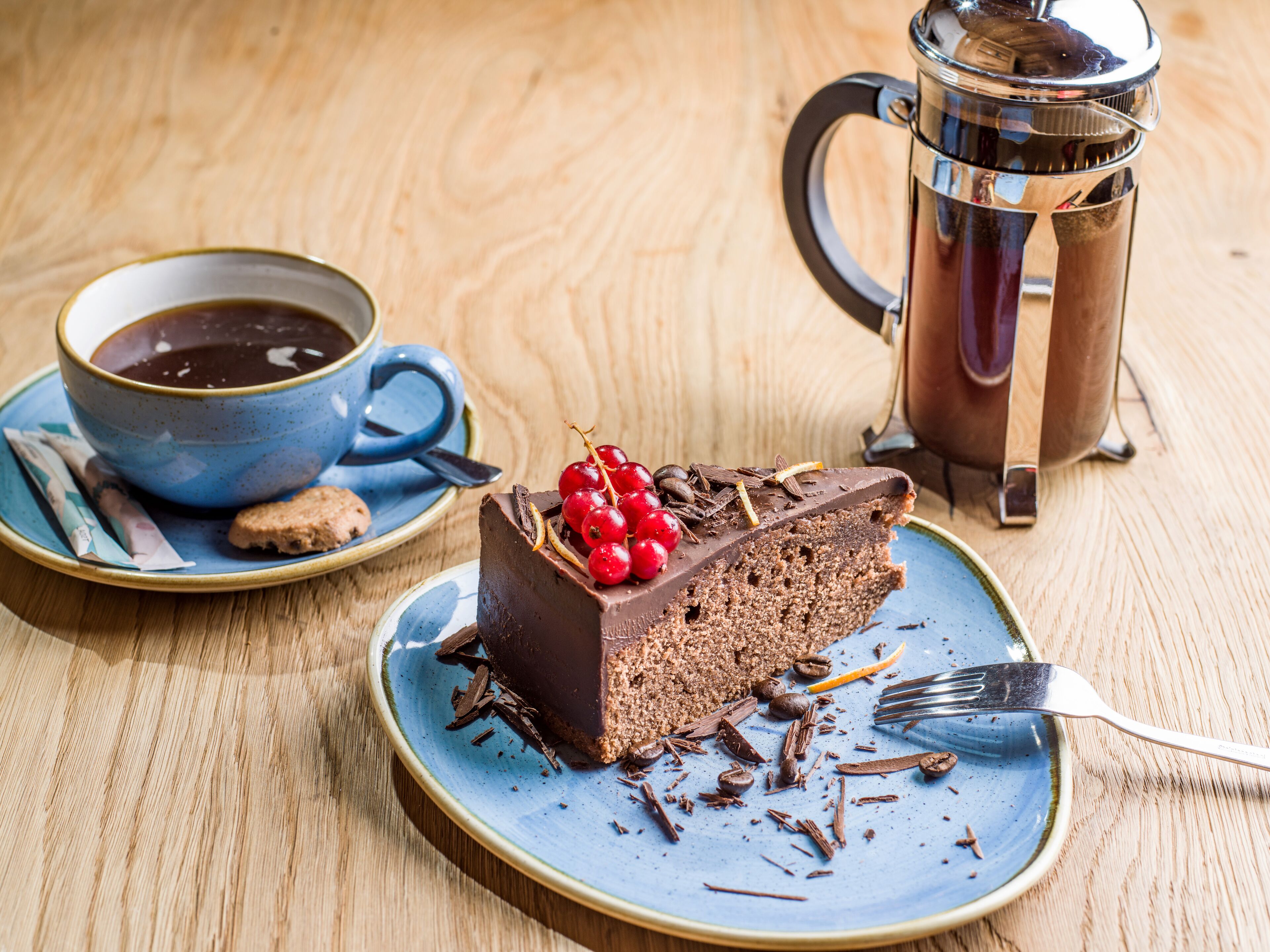 A slice of chocolate cake with berries on a plate, a cup of coffee, and a coffee pot on a wooden table.