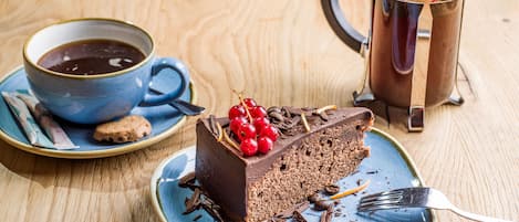 A slice of chocolate cake with berries on a plate, a cup of coffee, and a coffee pot on a wooden table.