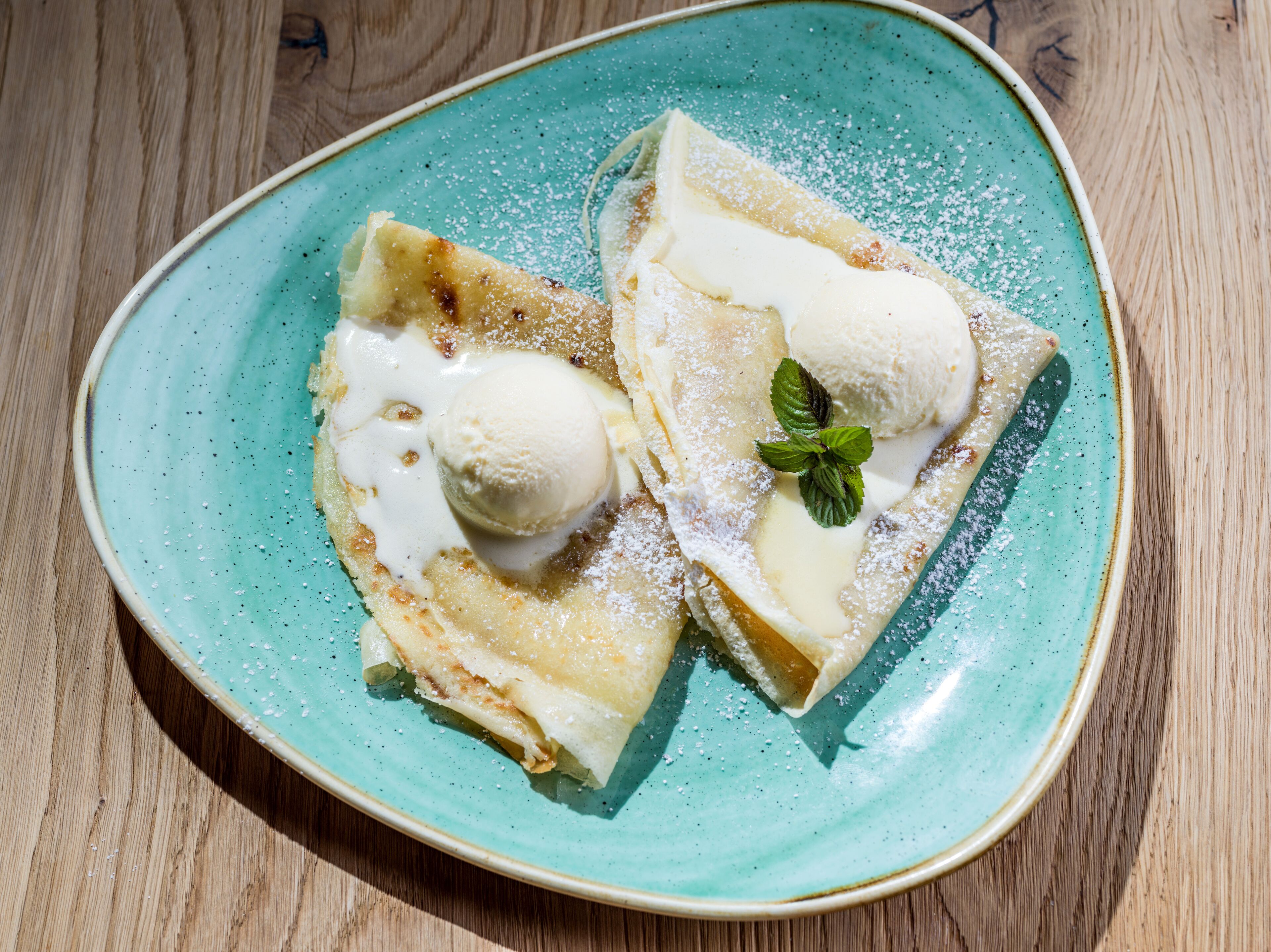 Ein Teller mit zwei Crêpes, belegt mit Vanilleeis und einem Minzblatt, bestäubt mit Puderzucker.