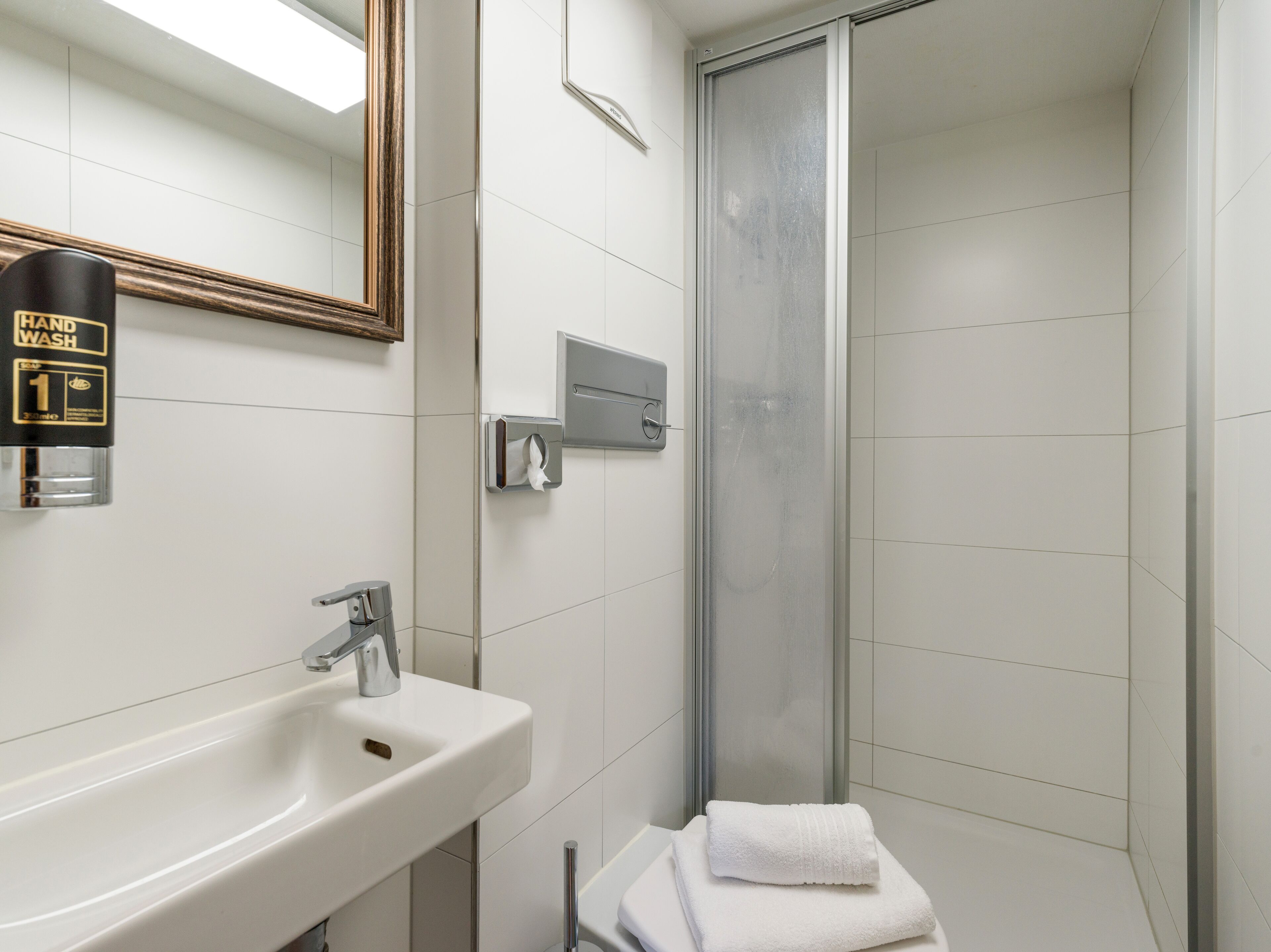 Un baño con lavamanos blanco, dispensador de jabón líquido, toallero y cabina de ducha con paredes de vidrio.