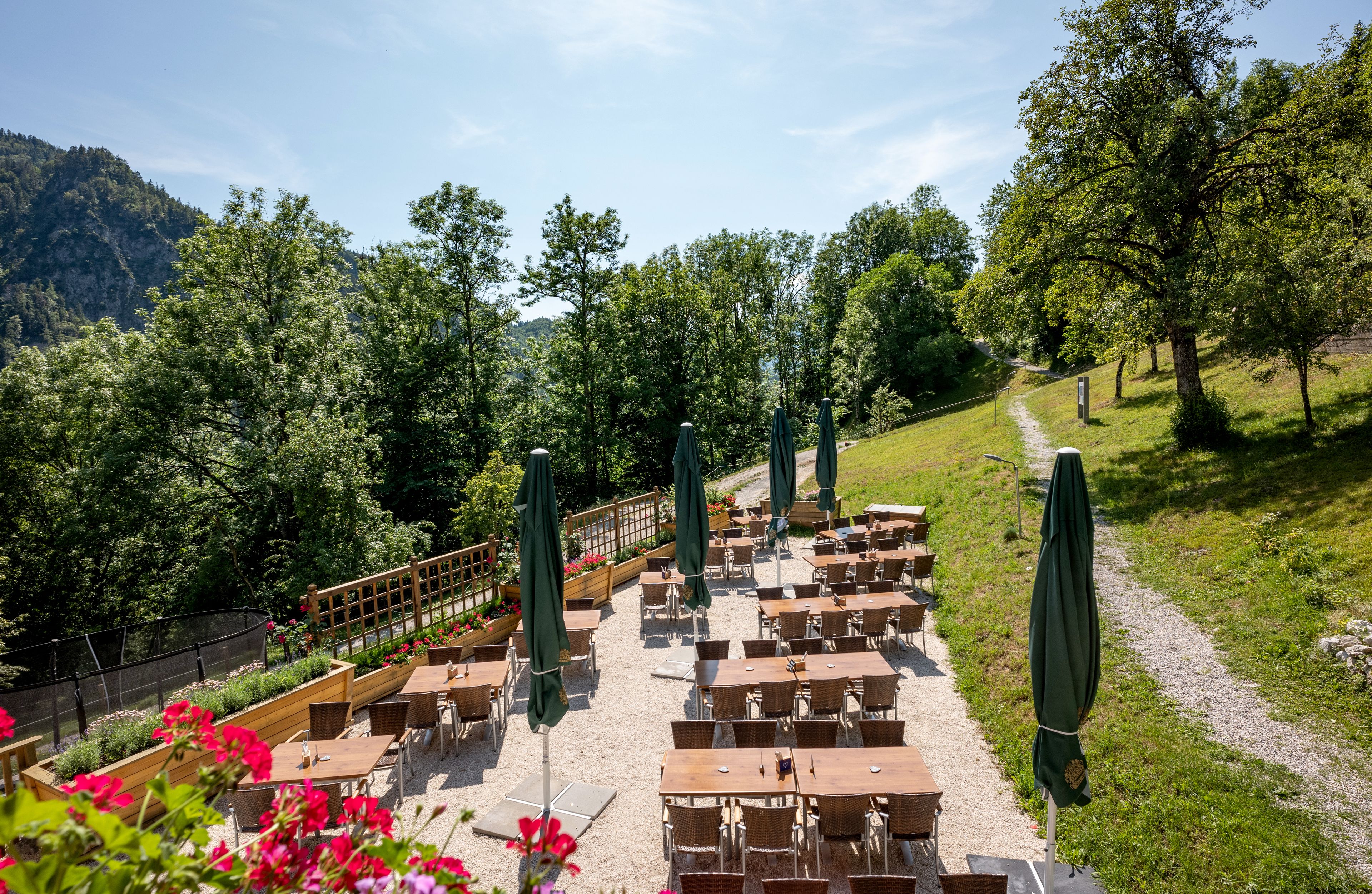 An outdoor seating area with tables and chairs, surrounded by greenery and a path leading to a mountain.
