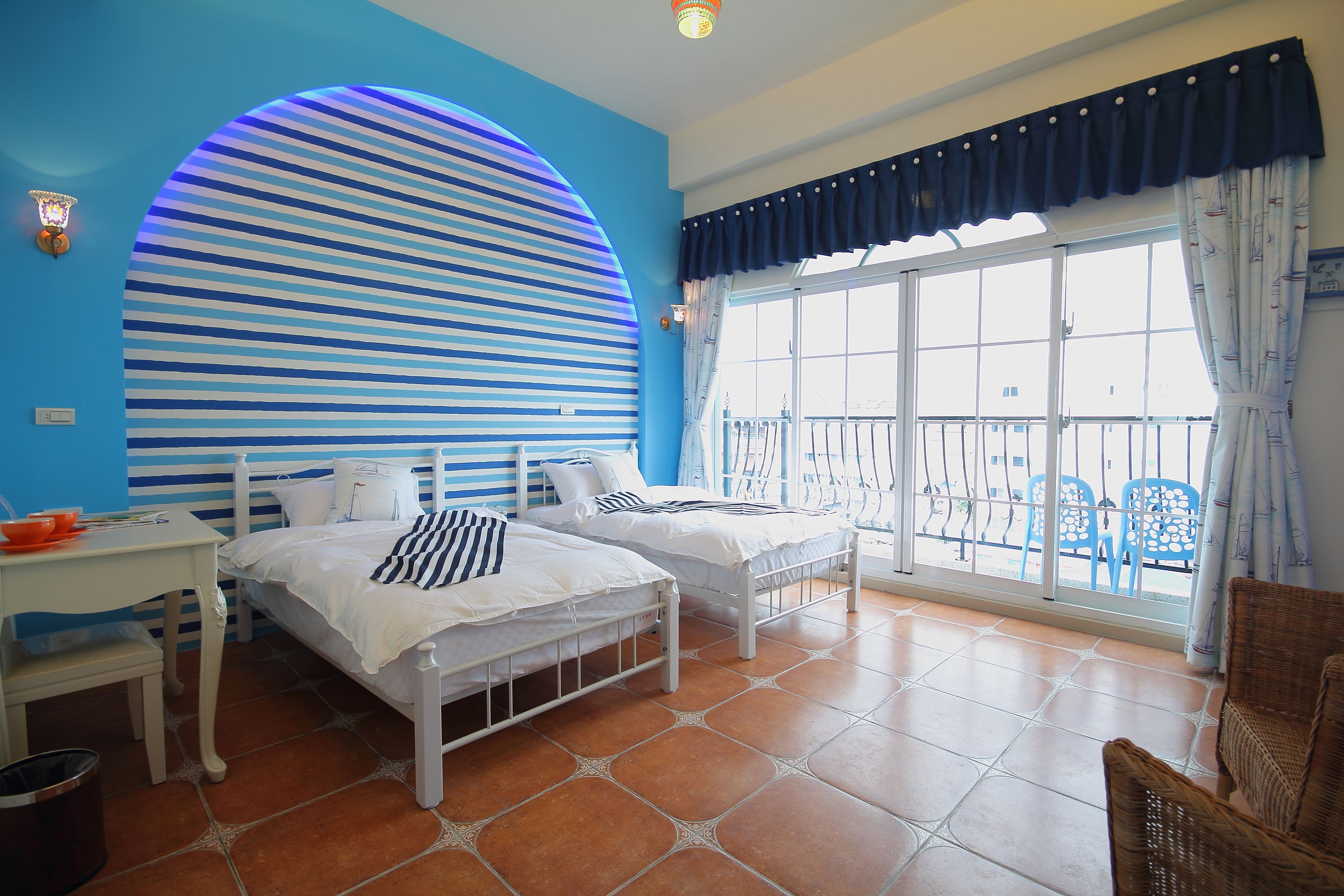 A room with two beds, a desk, and a chair. There is a large window with curtains and a view of the outside.