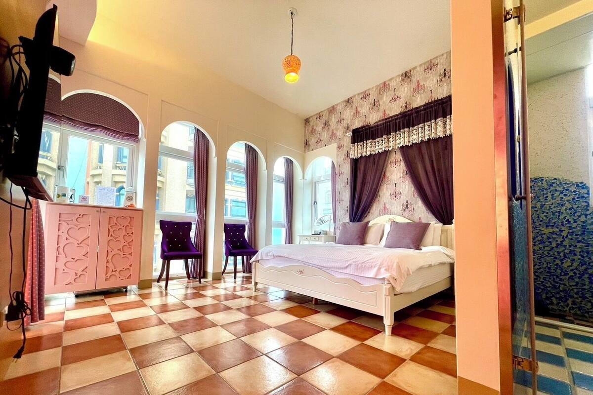 A hotel room with a checkered floor, a bed with a white headboard, purple curtains, and a view through arched windows.