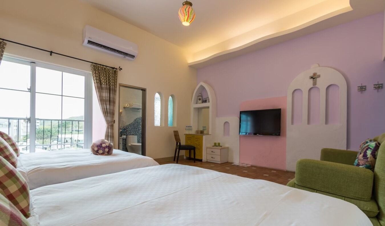 A hotel room with two beds, a TV, a small desk, a chair, and a balcony with a view.
