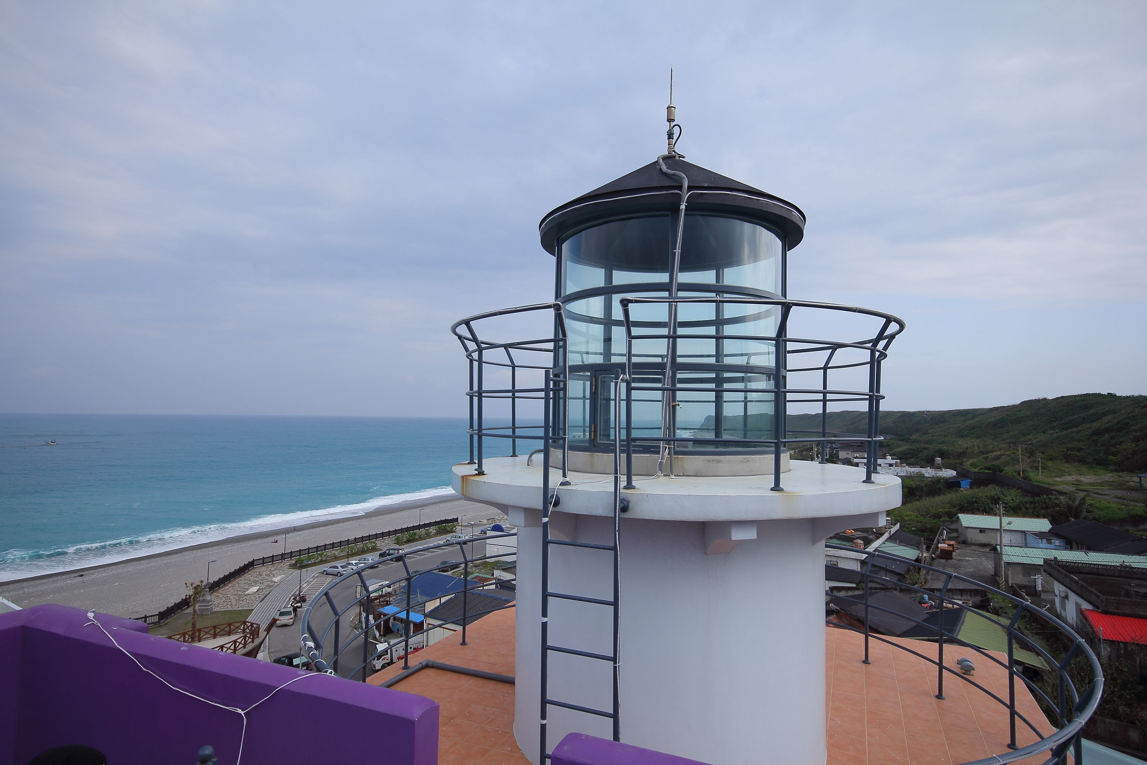 Un faro con cerramiento de cristal, situado en una terraza costera con vista al mar y colinas al fondo.