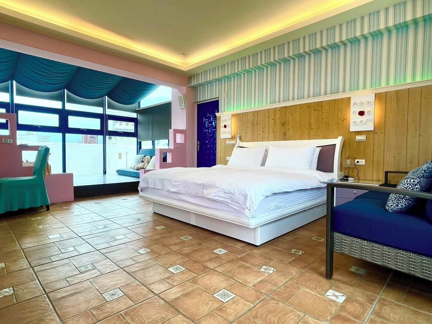 A modern hotel room with a large bed, a seating area with a blue sofa, a small table, and a view through large windows.